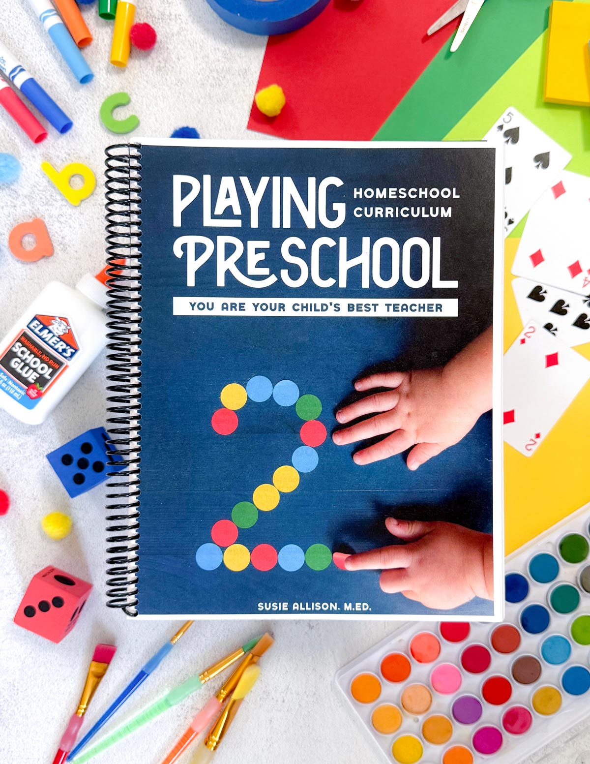 Playing Preschool homeschool curriculum spiral-bound book with a navy cover, lying on a white surface surrounded by crayons, scissors, glue, and a watercolor palette. The cover shows a child's hands next to the number 2 formed from colored dot stickers.