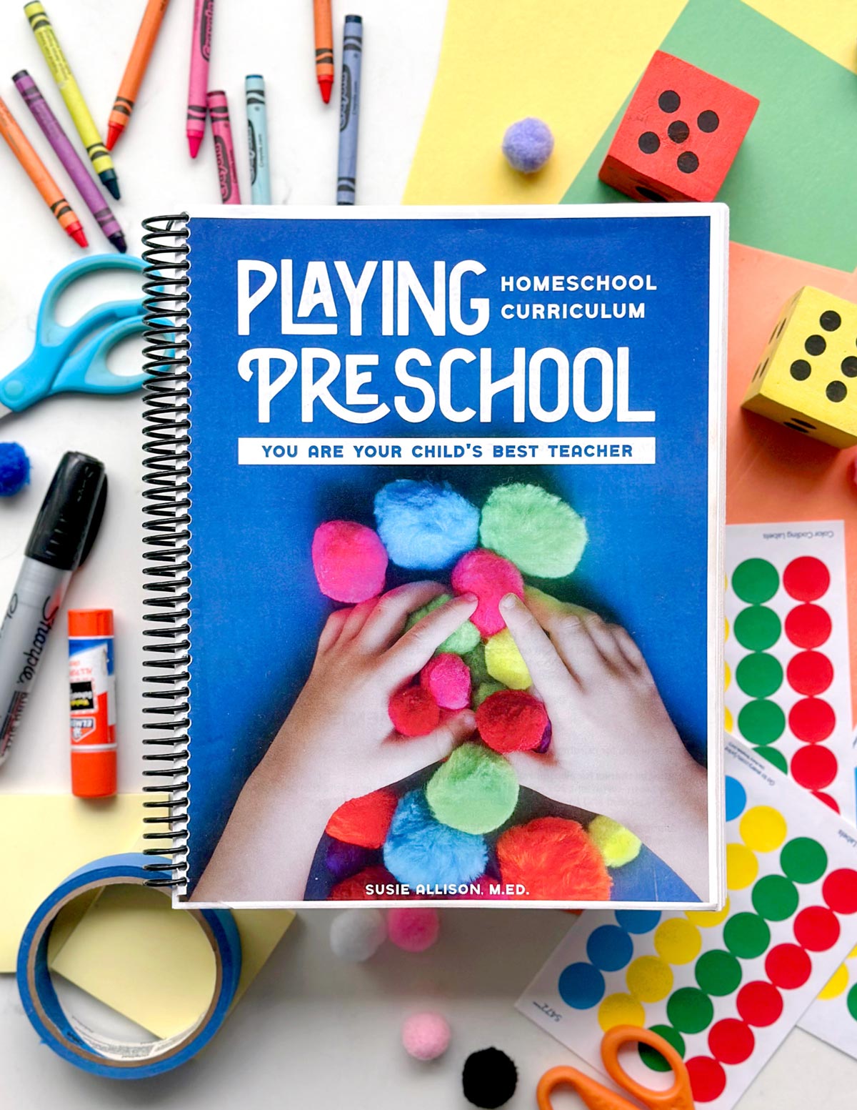 Playing Preschool homeschool curriculum spiral-bound book with a blue cover, lying on a white surface surrounded by crayons, scissors, glue, pom-poms, and colorful paper. The cover shows a child's hands nestled in a pile of colorful pom-poms.